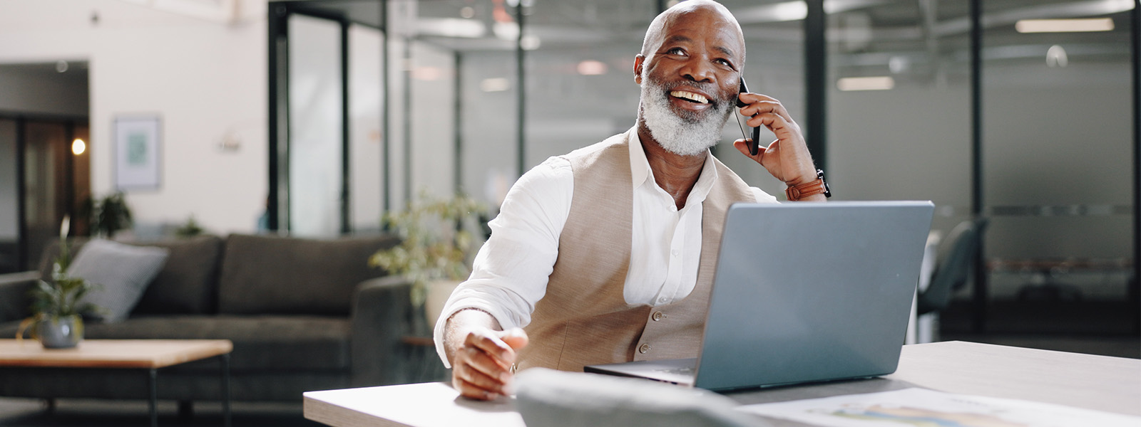 A sales professional with a white beard, holding a laptop in front, talking on a phone, and smiling.