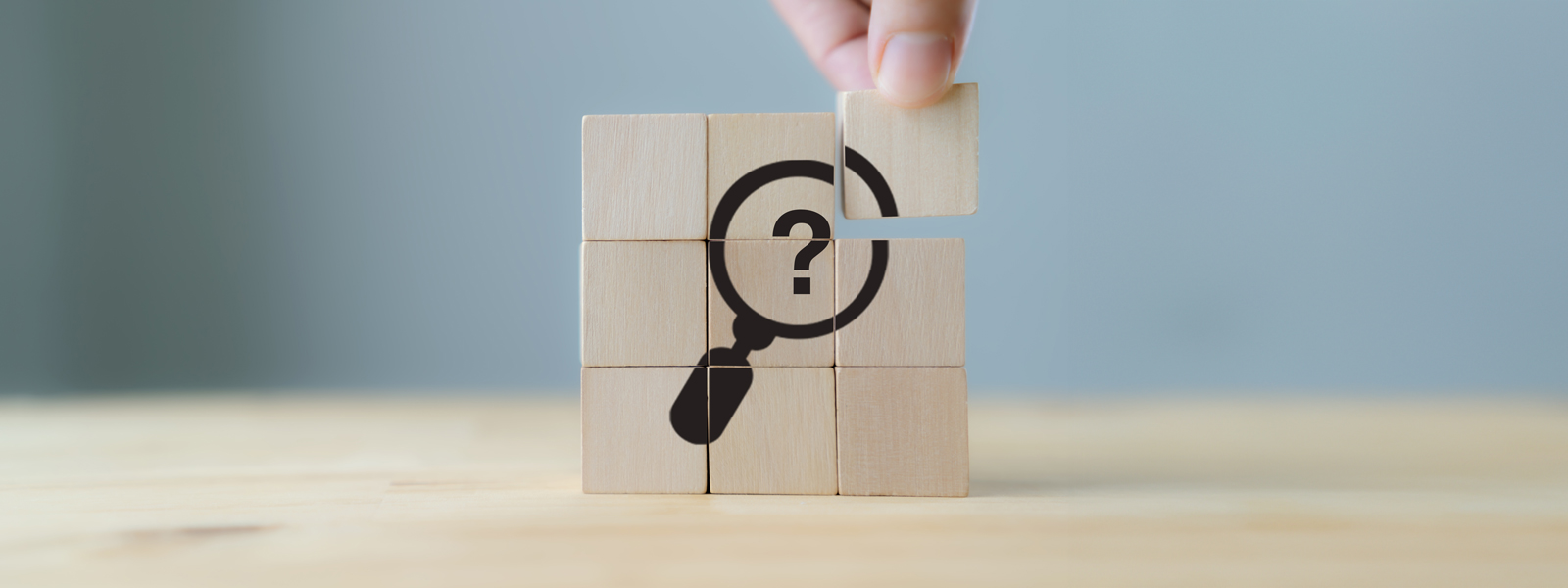 A close up photo of fingers assembling wood blocks forming an icon of a magnifying glass containing a question mark