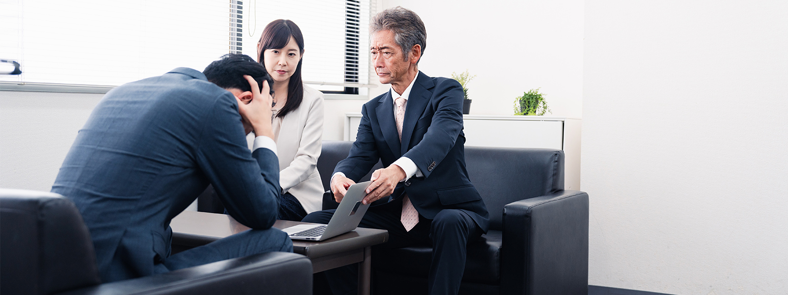 A man is holding his head while another person and a lady are showing something on a laptop.