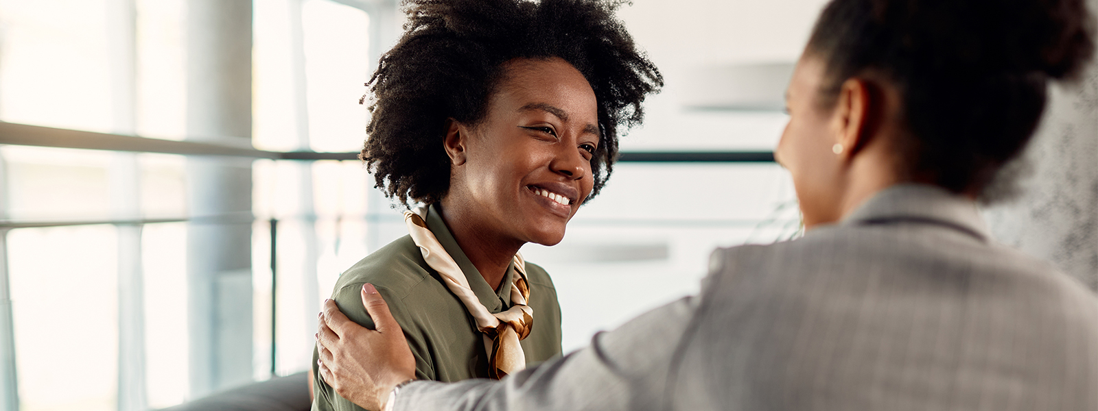 Two women warmly embracing in an office setting, one smiling broadly at the other